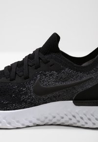 Zapatilla deportiva negra con un upper texturizado, con cordones negros y una suela de goma blanca lisa. Logo de Nike notable en negro en el lateral.