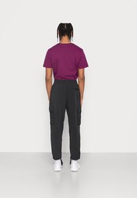 En person med flätat hår står vänd mot en slät ljus vägg, iklädd en lila kortärmad tröja, svarta cargobyxor och vita sneakers.