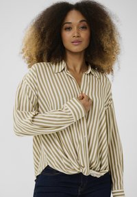 Camisa de botón a rayas en beige y blanco, con un diseño de nudo en la parte frontal y un ajuste relajado. Mangas largas y cuello clásico.