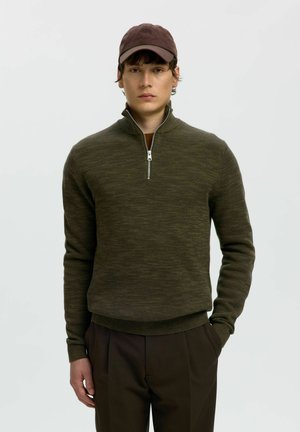 Jeune homme portant un pull à demi-fermeture éclair couleur olive foncé, une casquette marron et un pantalon sombre, debout avec une main dans la poche, sur un fond uni.