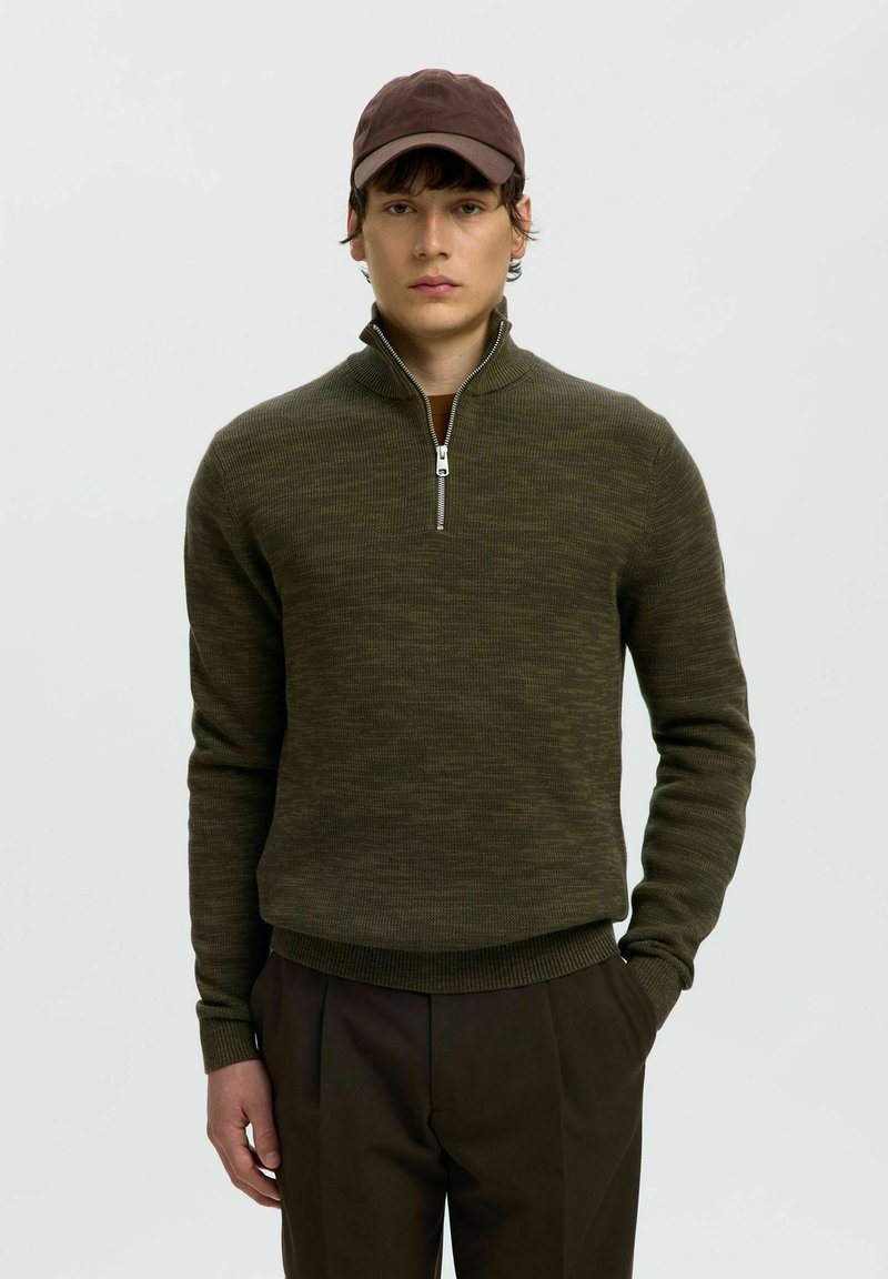 Jeune homme portant un pull à demi-fermeture éclair couleur olive foncé, une casquette marron et un pantalon sombre, debout avec une main dans la poche, sur un fond uni.