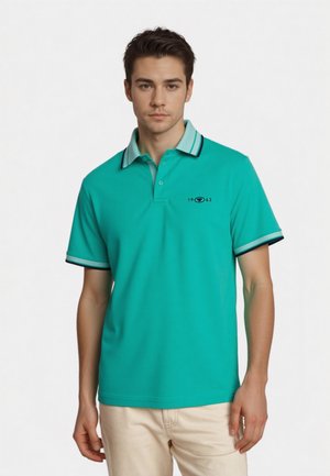 Young man wearing a teal short-sleeve polo shirt with light blue and black striped collar and sleeve edges, paired with beige pants.