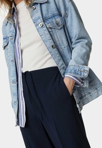 Ljusblå denimjacka med knäppta fickor, över en vit ribbad topp och marinblå byxor. Prydliga randiga manschetter från toppen är synliga.
