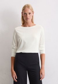 Weißes Langarmoberteil mit weitem Ausschnitt, weicher Textur, getragen zu hoch geschnittenen schwarzen Hosen. Einfaches, minimalistisches Design mit taillierten Ärmeln.