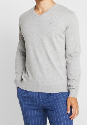 Homme portant un pull col en V gris clair à manches longues et un pantalon bleu à fines rayures, les mains détendues sur les côtés.