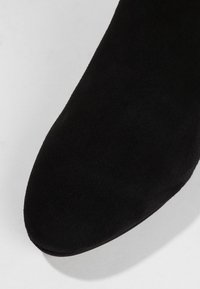 Botín de ante negro hasta el tobillo con punta redondeada y textura suave, que presenta un diseño minimalista sin herrajes o acentos visibles.