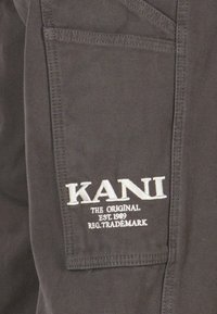 Pilkas audinys su matomomis siūlėmis ir užklijuota etikete "KANI" balta spalva, taip pat tekstu "ORIGINALAS EST. 1989 REG. PREKĖS ŽENKLAS".