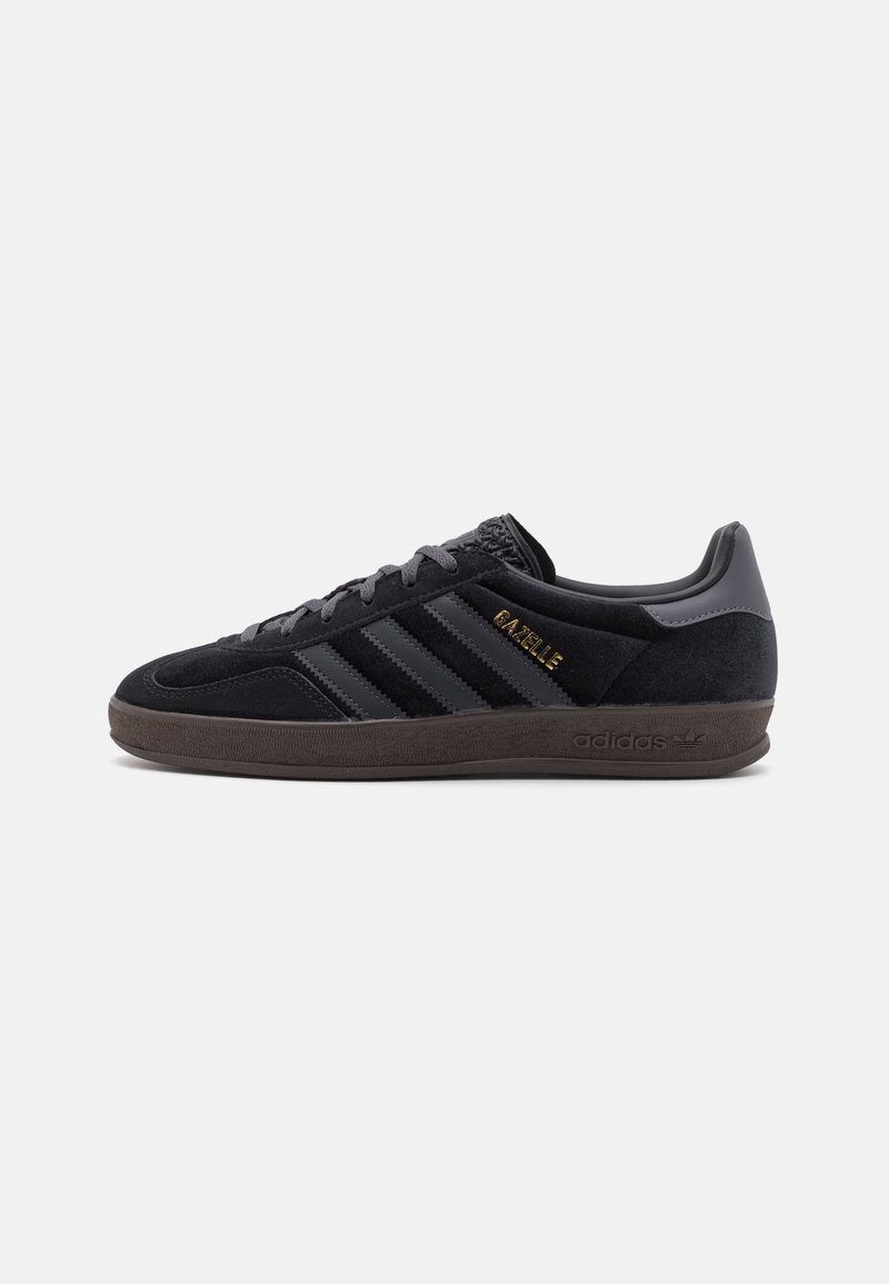 Baskets noires Adidas Gazelle avec tige en daim, trois bandes sur le côté, texte doré "GAZELLE" et semelle en caoutchouc marron.