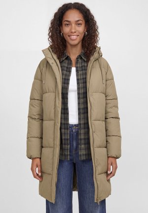 Une femme souriante porte un long manteau matelassé à capuche, couleur kaki, par-dessus une chemise à carreaux, un haut blanc et un jean bleu, sur un fond uni.