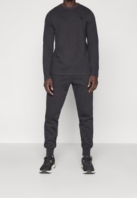 Schwarzes Langarm-Crewneck-Oberteil und passende Jogginghosen aus strukturiertem Material.getragen mit schwarzen Turnschuhen mit grünen Akzenten.