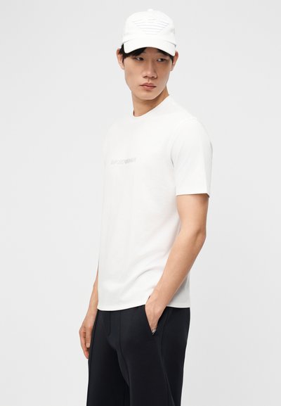 Νέος άνδρας που φοράει άσπρο καπέλο Armani και λευκό μπλουζάκι με το λογότυπο, και μαύρο παντελόνι, στέκεται με το ένα χέρι στην τσέπη και κοιτάζει προς τα πλάγια.