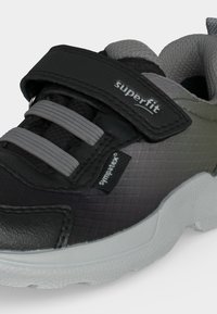 Gros plan d'une basket noire et grise avec des lacets élastiques, une bande auto-agrippante noire étiquetée "superfit," et une étiquette en tissu "sympatex".