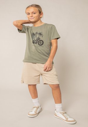 Garçon aux cheveux blonds, portant un t-shirt vert avec un motif de moto et un short beige, se tient avec une main derrière la nuque sur un fond uni.