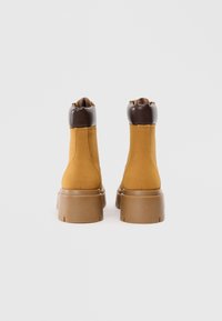 Ruskind, sennepsgule ankelstøvler med en mørkebrun læderkrave og en kraftig lys brun sål, der har minimale dekorative detaljer.