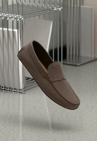 Brun læder loafersko med en tekstureret overflade, rund tå og syninger. Har et diskret logo som accent på siden.