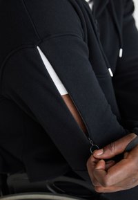 Crna zip up dukserica s bijelim detaljem na rukavu, sa bočnim zatvaračem i teksturiranom tkaninom. Ruka drži povlačenje zatvarača.