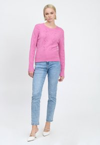 Giovane donna in piedi che indossa un maglione a maglia rosa, jeans azzurri chiari e mule bianche con tacco e cinturini di strass su uno sfondo bianco.