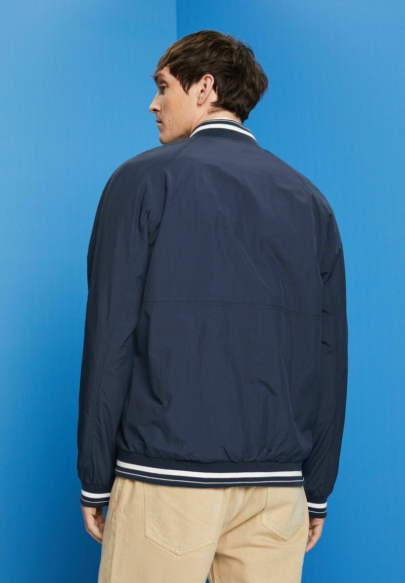 Veste légère marine avec col et poignets côtelés, présentant des rayures blanches et une coupe décontractée, associée à un pantalon de couleur claire.