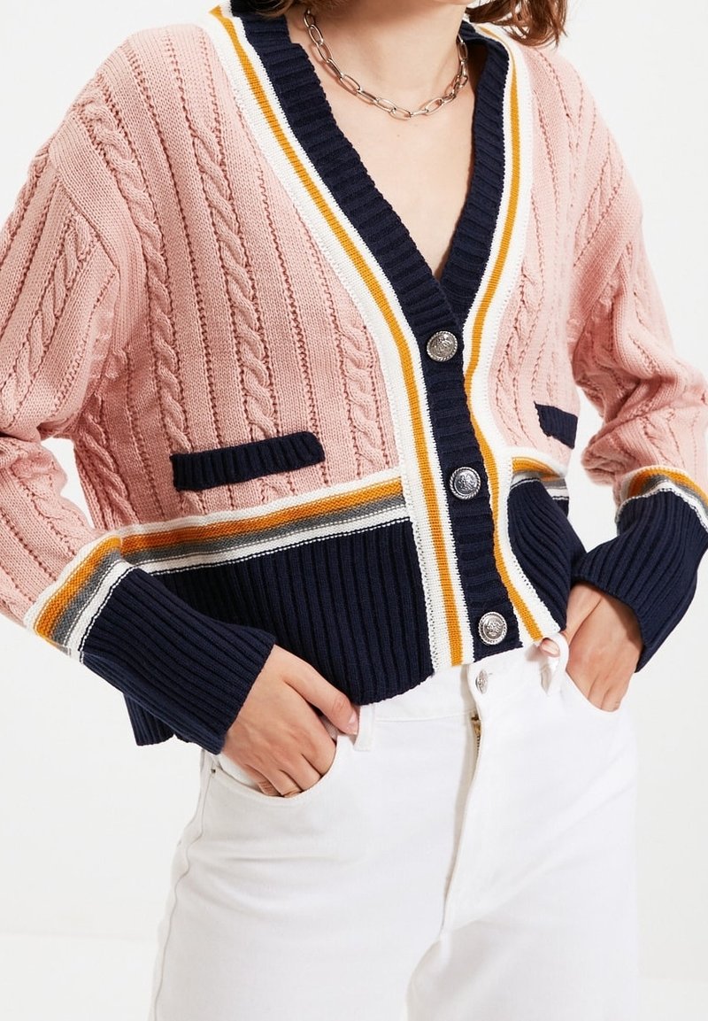 Femme portant un cardigan en maille torsadée rose avec un bord rayé bleu marine, blanc et jaune et des boutons argentés, associé à un pantalon blanc.