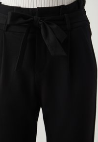 Black trousers featuring a tied bow at the waist, smooth texture, and pleated detail at the waistband. Flat front design.