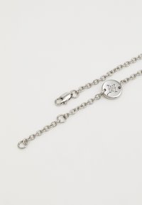 Pulseira de corrente de prata com elos ovais, um pingente redondo com iniciais estampadas e fecho de clasp. Textura suave por toda a parte.