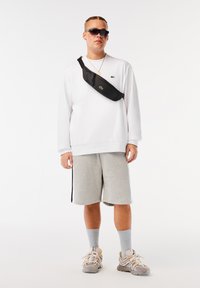 Schwarze Umhängetasche, quer über einem weißen Fleece-Sweatshirt getragen, kombiniert mit grauen Shorts und beigen Sneakers mit mehrfarbigen Akzenten.