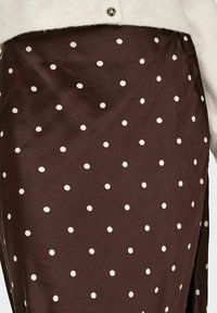 Jupe marron à pois blancs, en tissu lisse, avec un ourlet asymétrique et un léger éclat. Assortie à un cardigan léger.