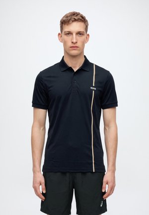 Un homme porte un polo noir BOSS orné d’une bande verticale dorée, assorti à un short noir, sur un fond clair uni.