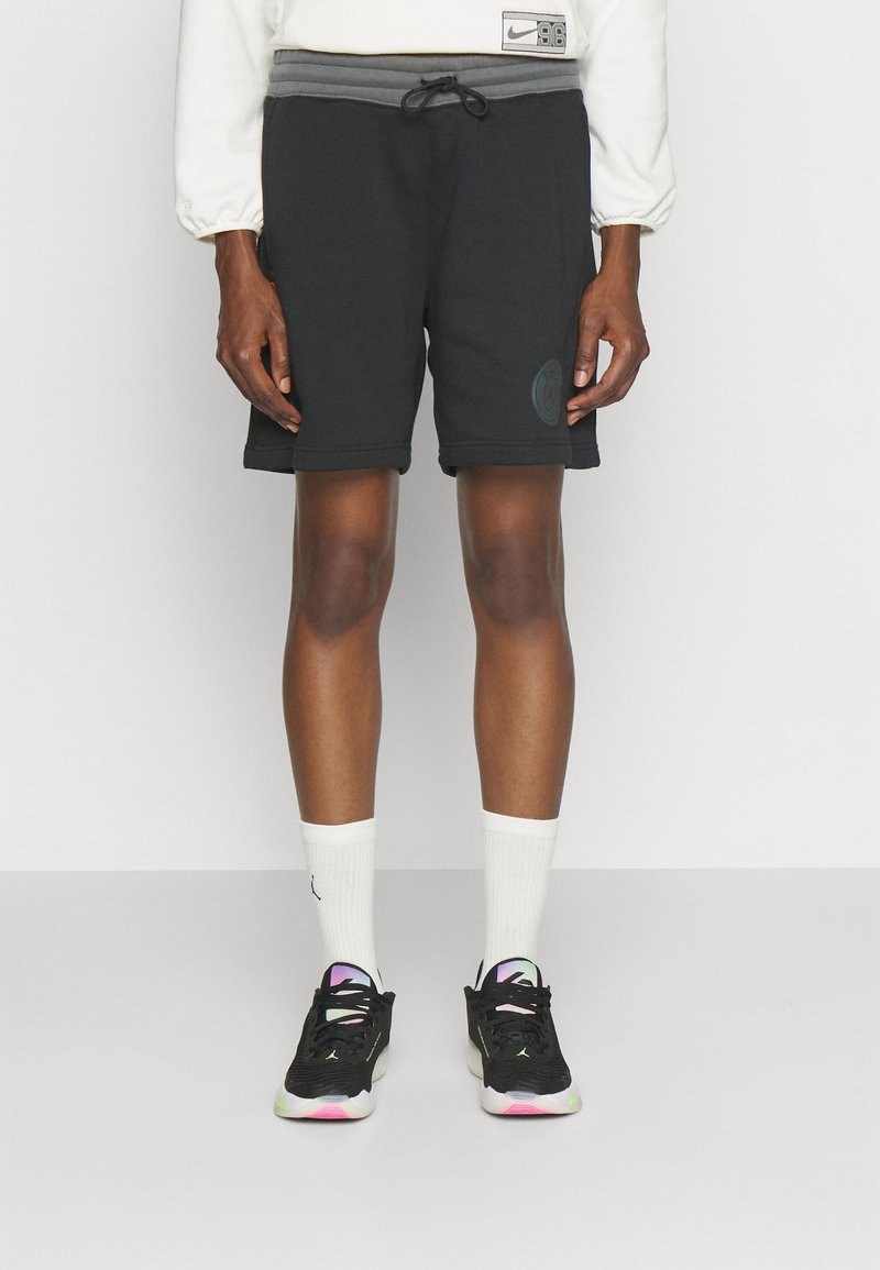 Osoba v černých šortkách, bílých ponožkách a černých teniskách s iridescentními akcenty stojící na neutrálním pozadí.