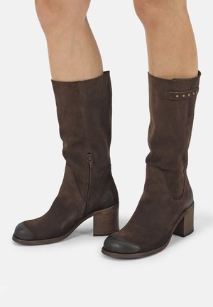Braune Wildleder-Stiefel bis zur Mitte der Wade mit Blockabsatz, seitlichem Reißverschluss und dekorativem Riemen mit Nieten, getragen an nackten Beinen vor einfarbigem Hintergrund.