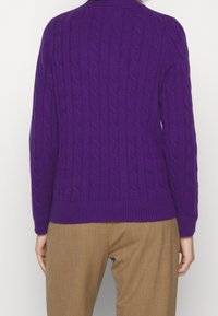 Lila kabelstickad tröja med ribbad nederkant och manschetter, med ett texturerat mönster. Matchas med ljusbruna byxor.