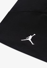 Μαύρο βαμβακερό ύφασμα με εμφανή υφή, που διαθέτει λευκό λογότυπο Jumpman. Ο σχεδιασμός είναι απλός, χωρίς μοτίβα ή επιπλέον διακοσμητικά στοιχεία.