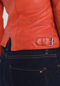 Kožená bunda v jasně oranžové barvě s podrobným prošíváním, přiléhavý design a nastavitelné přezky na boku. Spárovaná s tmavými džíny.