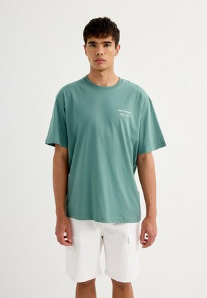 Camiseta de algodón verde con mangas cortas, cuello redondo y texto blanco "CONFIAR EN TU OBJETIVO" en el pecho. Se lleva con pantalones cortos cargo blancos.