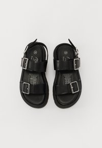 Sandálias de couro preto com duas fivelas ajustáveis, calcanhar fechado e sola texturizada, com um rótulo indicando o tamanho 28.