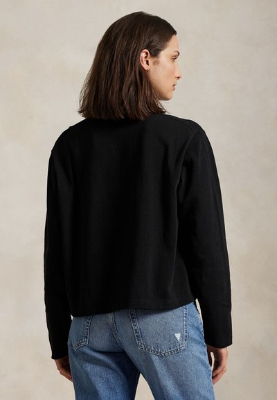 Nő vállig érő barna hajjal, fekete hosszú ujjú felsőt és kék farmert visel, háttal egy egyszínű háttér előtt.