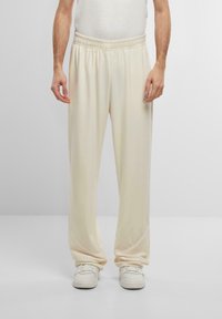 Pantalones de chándal color crema con cinturilla elástica y sutiles detalles de costura lateral. Combinados con zapatillas blancas. Textura suave en todo el pantón.