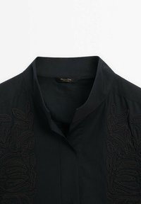 Col de chemise noir avec des motifs de feuilles brodées subtilement de chaque côté, présentant un col montant mandarin et une étiquette intérieure visible.