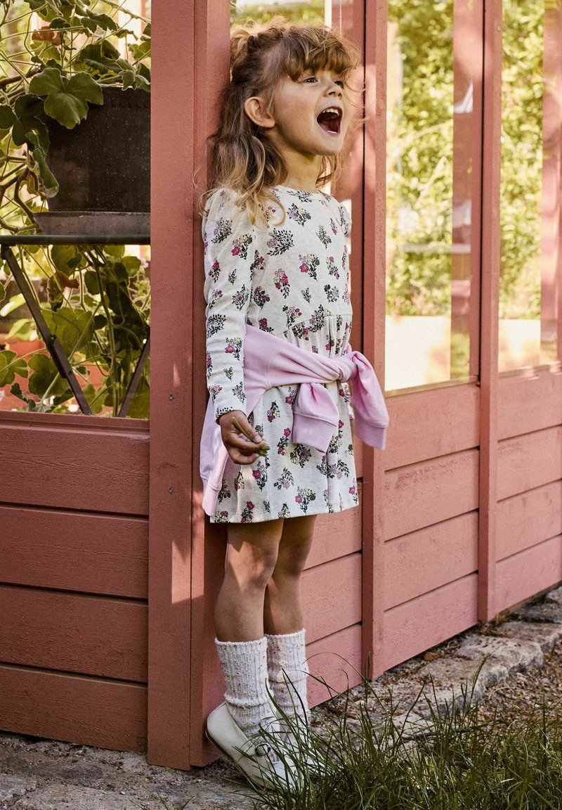 Jeune fille en robe fleurie avec un pull rose noué autour de la taille, debout et chantant à côté d'une structure en bois dans le jardin, avec des plantes à proximité.