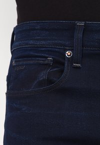 Mörkblå denimjeans med hög midja, subtil slitning, kontrastsöm och en klassisk femfickdesign med metallhardware.