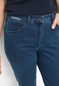 Jean en denim bleu foncé avec une texture lisse, présentant un design classique à cinq poches et une fermeture à bouton à la taille.