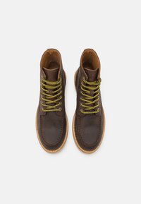 Botas de cuero marrón con cordones amarillos, punta redonda, con detalles de costura y una suela de color claro. Textura suave con un diseño estructurado.