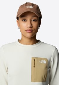 Gorra de béisbol marrón con logotipo, combinada con una camisa blanca texturizada que presenta un bolsillo marrón claro y un detalle de cordón.