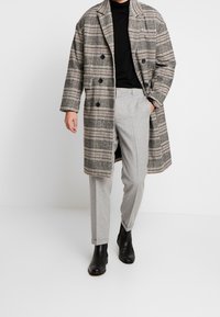 Geruite jas in grijstinten, rood en crème met dubbelgeknoopte knopen. Gecombineerd met lichtgrijze broek en zwarte enkelboots.