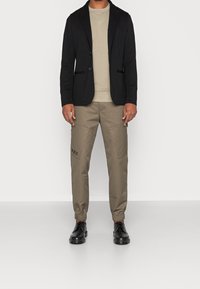 Svart blazer över en beige sweatshirt, parat med khaki joggerbyxor med sidofickor och svarta detaljer. Svarta skor avslutar outfiten.