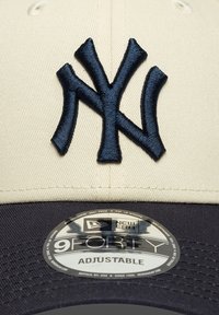 Boné cor creme com logótipo "NY" bordado em azul marinho. Design com viseira curva e tira ajustável. Apresenta uma etiqueta redonda com o logotipo "9FORTY".