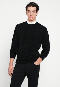 Μαύρο πουλόβερ με υφή σε σχέδιο, ριγέ μανσέτες και τελείωμα, στρογγυλή λαιμόκοψη, φορεμένο πάνω από ένα λευκό πουκάμισο με γιακά, με εφαρμοστή σχεδίαση.