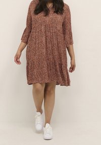 Robe rouille imprimée de fleurs, à design évasé, manches ¾ et ourlet à volants. Associée à des baskets blanches. Tissu léger.