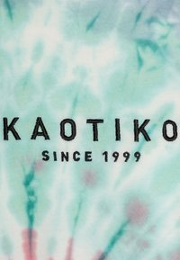 Jasnoniebieska tkanina tie-dye z czarnym haftowanym napisem "KAOTIKO" i "SINCE 1999" na miękkim, teksturowanym tle.