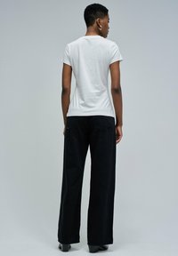 Mujer con el cabello corto, vestida con una camiseta blanca de manga corta ceñida, pantalones negros de pierna ancha y zapatos de tacón negros, de pie de espaldas.